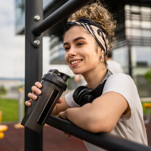 Junge Frau mit einem schwarzen Shaker, lächelnd am Geländer, sportlich gekleidet mit Kopfhörern.
