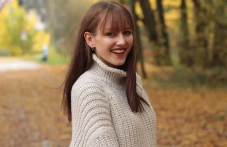 Frau mit braunen Haaren trägt einen hellen Pullover in der Natur, umgeben von herbstlichen Farben.