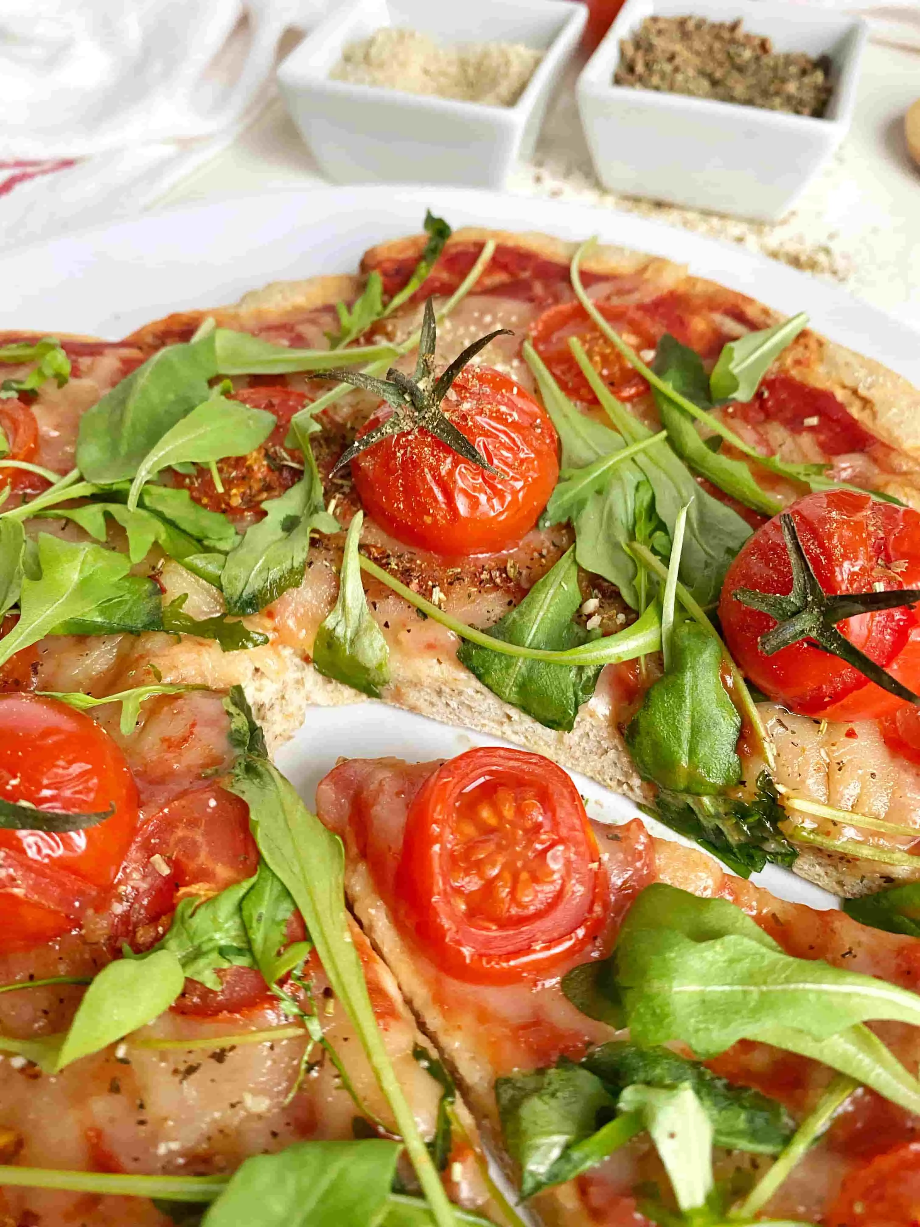 Leckere Protein-Pizzateig-Pizza mit Kirschtomaten, Rucola und Gewürzen auf weißem Teller.