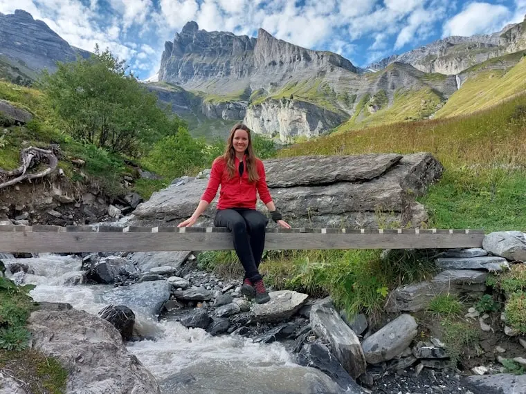 Frau in rotem Pullover sitzt auf einer Holzbrücke über einem Gebirgsbach, im Hintergrund beeindruckende Felsen.